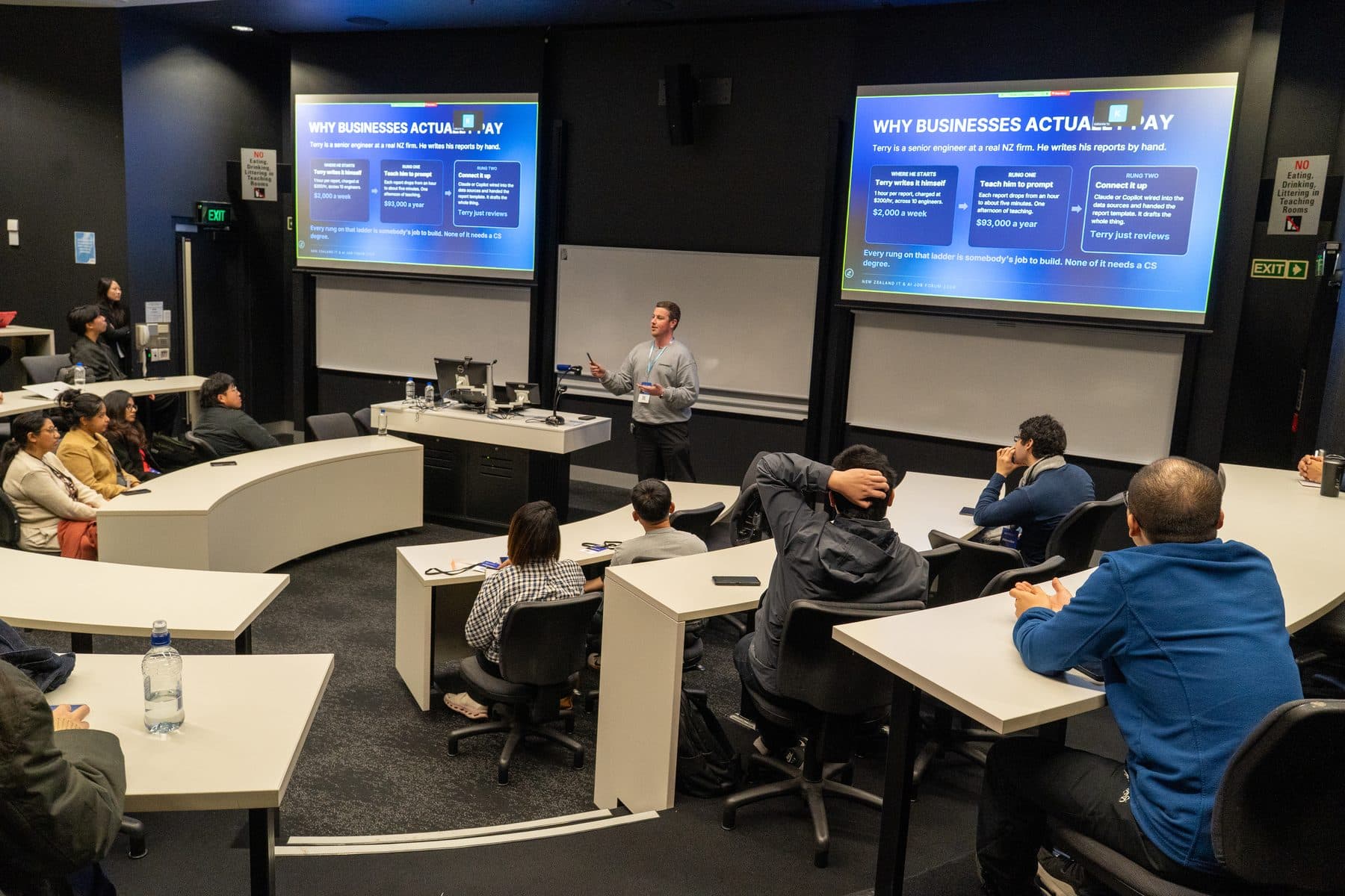 Wide view of a University of Auckland lecture theatre with Blake Harkness presenting to students between two large screens