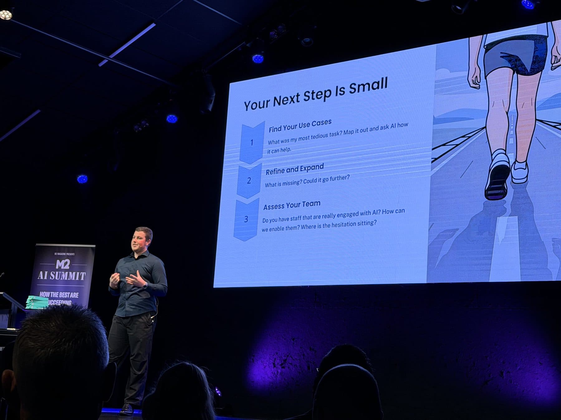 Blake Harkness on stage at the M2 AI Summit in front of a slide reading "Your next step is small"